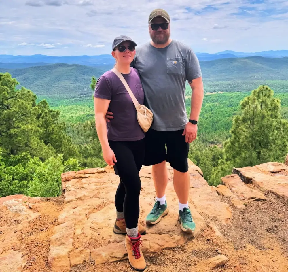 David and Dalis standing on a rocky cliff edge with a vast forested landscape and mountains in the background